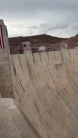 The video shows a wide, sweeping view of the Hoover Dam. The camera pans from left to right, showcasing the immense scale of the concrete structure. The dam's curved face is visible, with distinct horizontal lines indicating its construction. At the top of the dam, American flags are displayed on towers. Beyond the dam, a rugged, mountainous landscape with red-brown rocks is visible under a cloudy sky. Power lines and transmission towers can be seen in the distance. The video also captures a glimpse of the power plant facilities located at the base of the dam. The water level in the reservoir appears to be relatively low. There are no people visible in the video.