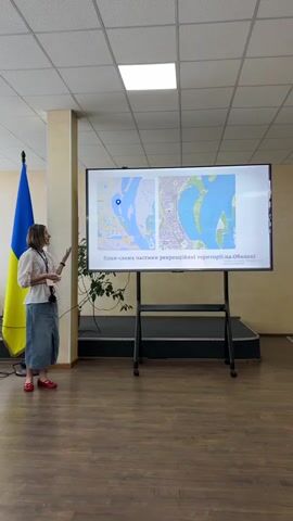 A woman is giving a presentation in what appears to be a conference room or lecture hall. She is standing to the left of a large screen displaying maps. The screen shows two aerial views of a river with islands, and text below in Ukrainian reads "План-схема частини рекреаційної території на Оболоні" which translates to "Schematic plan of part of the recreational area on Obolon". The woman is wearing a light-colored embroidered shirt, a denim skirt, and red shoes. She is gesturing with her hands as she speaks, and her expression suggests she is engaged in her presentation.

To the left of the woman is a Ukrainian flag. The room has a wooden floor and a drop ceiling with fluorescent lighting. There is a potted plant to the right of the screen. The overall scene suggests a formal or semi-formal educational or professional event. The lighting in the room is bright, and there are no windows visible, so the time of day and weather are not discernible from the image. The location is identified as Myronivka, Ukraine, however, the content on the screen refers to Obolon, which is a district in Kyiv, Ukraine, suggesting a potential discrepancy or that Obolon is being used as a geographical reference point.