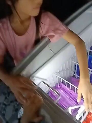 A young girl wearing a pink t-shirt is reaching into a chest freezer filled with ice cream. She is looking down into the freezer, her right hand is near the back of the freezer, and her left hand is on the edge of the freezer. The freezer contains several rows of ice cream bars, many of which are wrapped in purple and blue packaging. There are also some ice cream bars with red packaging visible at the bottom of the freezer. Another hand, presumably belonging to someone else, is visible in the foreground on the left side of the frame, also near the edge of the freezer. The lighting suggests an indoor setting, possibly a store or home. The girl appears to be choosing an ice cream.
