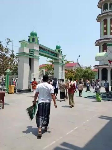 The video shows a large paved courtyard in front of a building with a gate. Several people are walking in various directions, including men, women, and children. Many appear to be dressed in traditional Muslim attire. The gate is a decorative arch structure painted white with green accents and small dome-shaped caps on top. The building in the background appears to be a mosque or religious building with multiple stories. Several trees are visible.  There are benches and a small cart or vehicle in the courtyard. The sky is clear and blue.