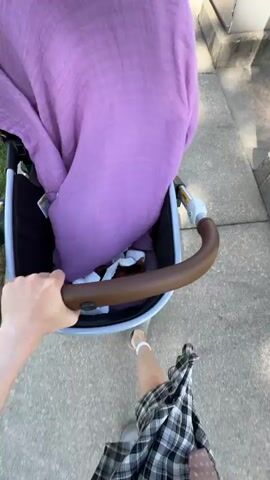 A person is pushing a stroller down a sidewalk. The stroller is covered with a purple cloth. The person is wearing a grey top and a plaid skirt. The person has short, red hair and is smiling. The person passes a building with a unique, curved roof. The sidewalk is next to a grassy lawn. The video is shot from the perspective of the person pushing the stroller.