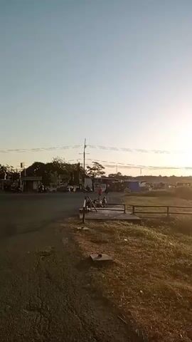 The video shows an outdoor scene at sunset. The sun is large and bright in the sky, casting a warm glow on the landscape. In the foreground, there is a grassy area next to a dirt path and a body of water. In the middle ground, there is a dirt road with some vehicles and people. In the background, there are trees and buildings. The overall impression is one of a peaceful, rural setting as the day comes to an end.