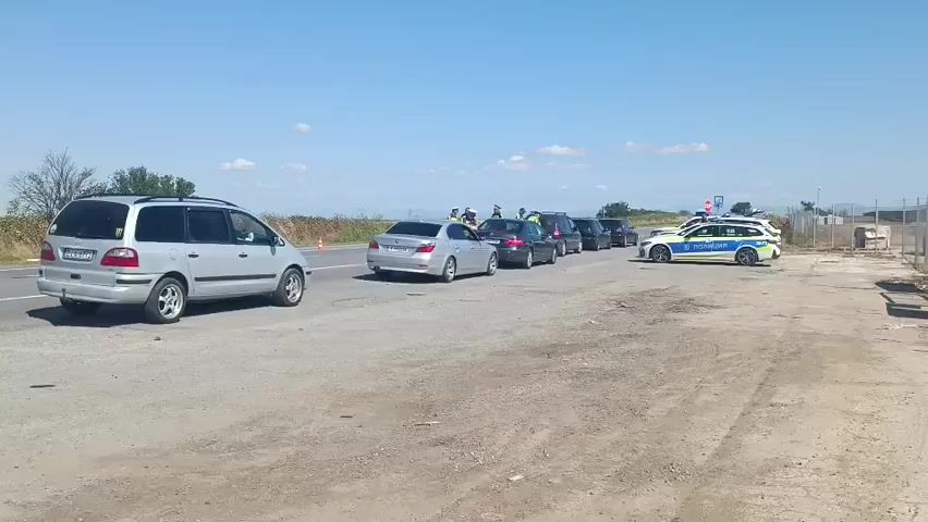 A line of vehicles is stopped on a rural road under a clear blue sky. Several people in uniform, likely police officers, are interacting with the drivers of the vehicles. Two marked police cars, one white with blue and yellow accents and the other a similar design, are present. The police cars are equipped with flashing lights. A silver minivan is positioned at the left side of the frame, facing away from the camera. Further down the road, several cars are lined up, including silver sedans and darker SUVs. The environment appears to be a flat, open area with sparse vegetation and a fence in the background. The road surface is asphalt, with a gravel shoulder. The overall scene suggests a traffic stop or checkpoint.