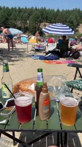 The video shows a close-up of a green table with drinks and snacks on it. In the background, people are enjoying a sunny day at a beach with trees and umbrellas. There are two drinks in plastic cups, one red and one amber, a bottle of green-capped beverage, and a bottle of what appears to be brown liquid. There's also a spray bottle with a gold cap and a label that says "DAX". Next to these items, a plastic bag filled with snacks, and a large loaf of bread are visible. The table is set on a sandy surface, and in the distance, several people are scattered on the beach, some under umbrellas and some in the water. The sky is clear and blue, indicating good weather.