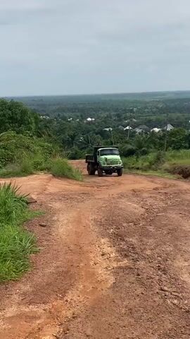 A light green dump truck drives on a dirt road. The road is bumpy and has some ruts. The truck is heading away from the camera. The truck has a black tarp covering the bed. In the background, there is a green, hilly landscape with trees and some houses. The sky is overcast.