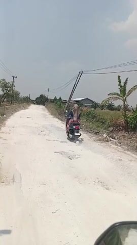 A motorcyclist rides down a dirt road. The road is unpaved and appears to be in a rural area. There are trees and houses on either side of the road. The sky is clear and the sun is shining. The motorcyclist is wearing a helmet and a jacket. The motorcycle is a scooter. The motorcyclist is carrying a backpack. The motorcyclist is looking ahead.