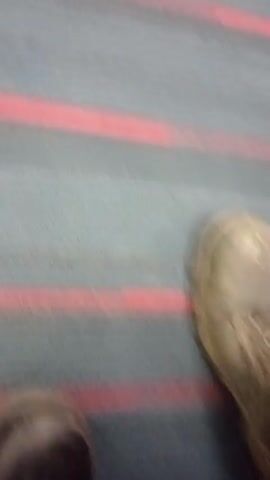 The video shows a view of a carpet with a horizontal pattern of gray stripes and red lines. A pair of dirty work boots is visible at the start of the video, followed by white tennis shoes. The camera perspective is low, suggesting the video was taken near the ground. The camera moves slightly, shifting the focus on the carpet pattern and the footwear passing by.