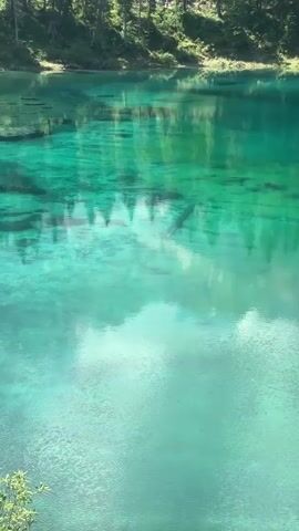The video displays a panoramic view of a vibrant turquoise lake surrounded by lush green trees and majestic mountains. The water is incredibly clear, allowing glimpses of submerged logs and the lakebed. Sunlight filters through the clouds, creating a dappled effect on the water's surface, which reflects the surrounding trees and sky. The camera pans slowly, capturing the serene beauty of the natural landscape. The scene is tranquil and picturesque, suggesting a peaceful, remote location.