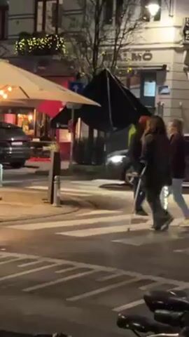 The video shows two people and another figure walking across a zebra-striped crosswalk in a European city at night. One of the individuals uses crutches to walk. The other person walks next to her, and another person follows behind. There is a car and a bicycle partially visible in the foreground, and buildings with illuminated lights are visible in the background. A restaurant with tables is visible to the left. The street and crosswalk are marked with white lines.