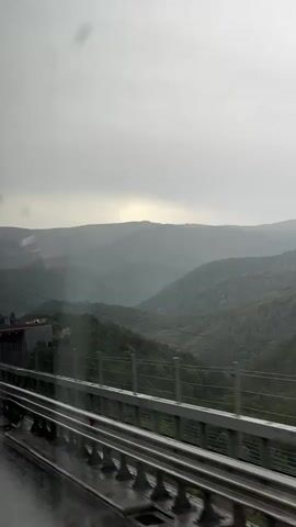 The video shows a view from a moving vehicle, likely a train, looking out at a mountainous landscape. The sky is overcast and appears to be raining, with visible streaks of rain falling. In the foreground, a metal barrier or railing runs horizontally across the frame, with a wet surface beneath it, indicating rain. Beyond the railing, green, tree-covered mountains rise and fall, with layers of mist or clouds obscuring the further peaks. A few buildings with red roofs are visible on the left side of the frame, nestled on a hillside. The overall impression is of a damp, moody, and scenic environment.