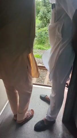 Two individuals, possibly men, are seen from the waist down. They are standing inside what appears to be a train car, indicated by the visible train tracks and landscape outside the window. Both are wearing traditional South Asian clothing. The person on the left is wearing a cream-colored kurta and salwar, with brown sandals. The person on the right is wearing a white kurta and salwar, with black slip-on shoes. The person on the left appears to be adjusting something above their head, possibly a turban or a piece of clothing. The person on the right is standing facing away from the camera, with their hands in their pockets. The background shows a green, rural landscape with trees and fields, suggesting the train is in motion and traveling through the countryside. The lighting is natural, indicating daylight. The overall scene captures a moment of travel within a train.
