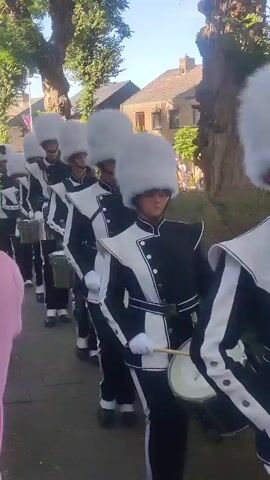 A group of people in uniform are marching and playing drums. They are wearing black and white uniforms with large white fluffy hats. The group is walking down a paved path with trees on either side. Houses are visible in the background. The sun is shining.