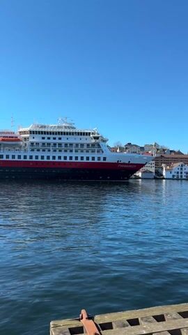 The video shows a large cruise ship, the Finnmarken of the Hurtigruten line, docked in a harbor. The ship is painted in black, red, and white. The water is calm and blue, and the sky is clear and bright. The city skyline is visible in the background. A tugboat is docked behind the Finnmarken. The camera is stationary. There are no people visible in the video.