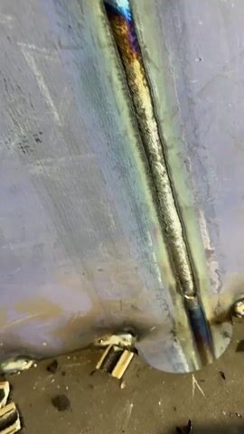 The video shows a close-up view of a freshly welded metal seam. The weld appears to be a continuous bead, with some discoloration indicating heat exposure, transitioning from blues and purples to yellows and whites. The metal surface around the weld is a dull grey with some subtle iridescent sheens. Along the bottom edge of the frame, small, rectangular wooden blocks are scattered, along with some dark debris. There are also several smaller, tack welds visible along the bottom edge, appearing as smaller, rougher blobs of metal. The overall impression is of a work-in-progress in a metal fabrication or repair setting.