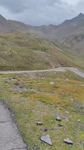 A white vehicle drives along a winding mountain road. The road is asphalt and curves sharply. The surrounding environment is mountainous with green grass and rocks. The sky is cloudy.