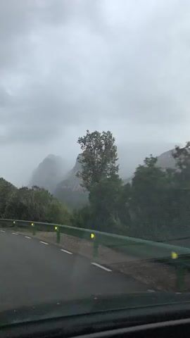 A car travels along an asphalt road with a green guardrail. White road lines are visible. The sky is overcast. Green foliage and trees obscure the view of mountain peaks in the distance. The overall impression is gloomy and wet. The camera is mounted inside the moving vehicle. Music plays.