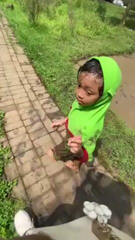 A child in a green head covering walks along a brick path in a grassy outdoor area. The child is barefoot and has mud on their clothing. The child looks up and briefly holds something in their hand before walking down the path. An adult reaches out a hand to the child as they walk. Other people are seen walking in the background. The environment includes grass, bushes, trees, and a muddy area. The weather appears sunny.