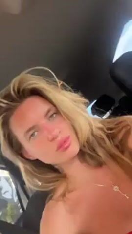 A fair-skinned, blonde woman sits in a car. She has a slight sunburn and her lips are glossy. She wears a silver necklace with a key-shaped pendant and her hair is tousled. She looks around and poses for the camera. A black car seat headrest is visible behind her and the sky can be seen through the car window.