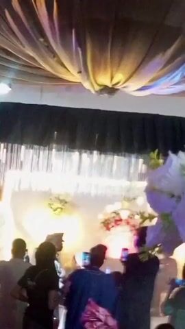The video shows a dimly lit indoor event, possibly a wedding reception or a formal gathering. Several people are visible, some dressed in traditional African attire. In the background, there is a decorated stage area with lighting, including string lights and spotlights. Floral arrangements and fabric drapes are part of the decor. Some individuals are standing and interacting, while others are seated. One person in the center appears to be dancing or moving to music. The camera work is shaky, suggesting it was filmed from a handheld device. The overall atmosphere appears festive.