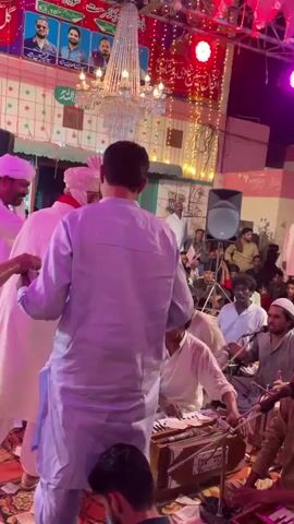 The video captures a lively indoor gathering, possibly a religious or cultural event. The scene is decorated with colorful banners featuring portraits and calligraphy, and adorned with sparkling chandeliers and string lights. A group of men in traditional attire are actively participating, with some dancing and others engaged in musical performance. One individual is playing a harmonium, a traditional Indian subcontinent instrument, while others watch and participate. The audience is seated on the floor, observing the proceedings. The atmosphere appears festive and energetic, with a sense of community and shared celebration.