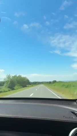 The video shows a view from inside a car looking out the windshield at a straight, paved road stretching into the distance. The sky is a clear blue with scattered white clouds. On either side of the road are green fields and trees. The dashboard of the car is visible in the foreground. The shot is steady, suggesting the car is in motion.