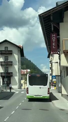 This is a rear-view, first-person perspective shot of a white and green bus driving down a street in Zell am See, Austria. The bus has the number "680" displayed on its back. The street is paved, with white lines marking the lanes and a pedestrian crossing. On the left side of the street, there is a multi-story building with balconies and windows. A lone person in a white shirt and dark pants walks away from the camera on the sidewalk. On the right side of the street, another multi-story building with balconies is visible, featuring a red "HOTEL" sign with white lettering. There are also traffic signs and construction barriers.

In the background, a lush green mountain rises, partially obscured by trees. The sky is a bright blue with large, fluffy white clouds, suggesting it is daytime and the weather is fair. The architecture of the buildings, the signage, and the mountainous backdrop strongly indicate a European alpine setting. The overall mood is calm and suggests a leisurely drive through a picturesque town. The visible text includes "HOTEL" on the sign and "680" on the bus. There is also text on the bus that reads "Salzburg Verkehr."