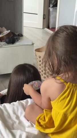 This video, filmed indoors, shows a young girl in a bright yellow dress brushing the hair of an unseen individual, likely an adult, who is lying on a white sheet. The girl has light brown hair and is focused on the task, holding a colorful hairbrush. The other person's dark brown hair is visible, indicating they are lying face down. The scene appears to be a casual, everyday moment in a home environment.

In the background, a light-colored wooden floor is visible, along with a beige laundry basket with a textured, circular pattern. There's also a white dresser or cabinet on the left. The room seems well-lit, suggesting it might be daytime. The sound of someone speaking in Russian can be heard in the background. The video has no visible text.