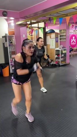 The video shows a group of women participating in a fitness class, likely a dance or cardio workout, in a gym. The primary focus is on a woman in the foreground wearing a black short-sleeved top and patterned pink shorts, actively engaged in punching and kicking motions. Her hair is pulled back in a ponytail, and she is wearing sneakers. To her left and slightly behind, another woman in a black top and dark shorts is performing similar movements. Further back, other women are visible participating in the class. The gym environment is characterized by pink walls, a dark floor with a grid pattern, and colorful banners strung across the ceiling. Various fitness equipment and shelves with products are visible in the background. The lighting appears to be artificial.