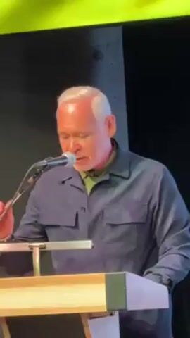 A man with short gray hair is speaking into a microphone while standing behind a wooden podium. He is wearing a dark blue collared shirt over a green shirt. He glances down at the podium. There is a bright light overhead.