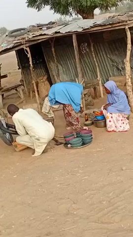 The video depicts a scene of everyday life in a rural setting. Three men are present, one dressed in a pink patterned outfit, another in brown, and the third kneeling and wearing white. The kneeling man appears to be working on the front tire of a motorcycle. Two women, both wearing blue hijabs, are seated nearby, seemingly preparing food. There are several colorful bowls on a tray next to them. A makeshift shelter constructed of corrugated metal and wooden posts stands in the background. The ground is sandy and bare. The general atmosphere suggests a modest, communal environment.