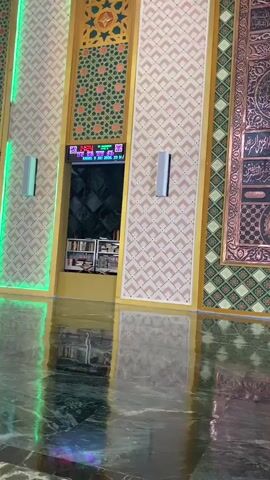 The video shows a panning view of the interior of a building, likely a mosque, with ornate decorations. The walls are covered in intricate patterns of gold, green, and beige. There are large, arched doorways with a green light strip outlining one of them. In the center of the frame, a digital clock displays the time and date. Below the clock, a shelf holds several books, suggesting a library or reading area. Two modern silver speakers are mounted on the wall. The floor is highly reflective, showing the lights and decorations above. The overall impression is one of opulence and spiritual significance.