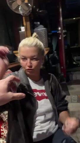 A person with blonde hair tied up is sitting in a dimly lit room. They are wearing a gray zip-up jacket over a white t-shirt with red writing. The person is holding their arm up to their face, covering their nose and mouth with their forearm. Their eyes are closed or looking down. In the background, there is a fan mounted on the ceiling, shelving with objects, and dark curtains. The person is sitting on a sofa with patterned cushions.