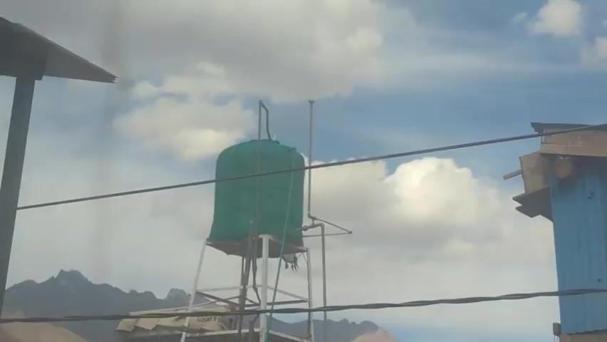 The video shows an outdoor scene with a water tank on a metal stand. The water tank is green and covered with a fabric shroud. There are clouds in the sky and mountains in the background. There are also some wires in the foreground. A small blue structure is visible on the right side of the frame. The scene appears to be in a dry, possibly arid environment.