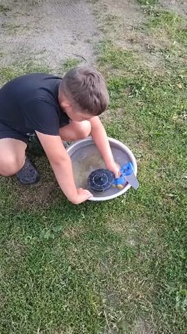 A young boy in a black t-shirt and shorts kneels on the grass, holding a blue and yellow toy water sprinkler with a black circular base in a metal bowl of water. The boy is manipulating the sprinkler, causing water to spray. A baby in a white and blue striped shirt and dark shoes walks towards the bowl of water. The scene takes place outdoors on a grassy area with some dirt.