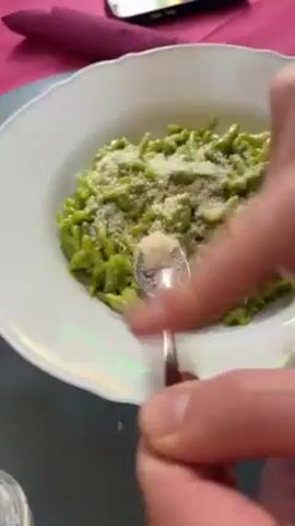 The video shows a close-up of a plate of green pasta being topped with shredded cheese. The pasta is in a white bowl with a patterned rim. A person is using a small spoon to sprinkle cheese onto the pasta. In the background, there is a pink tablecloth, a phone, a second plate of tomato-based pasta and glasses of red wine. The camera zooms in slightly and then pans to show more of the table setting.