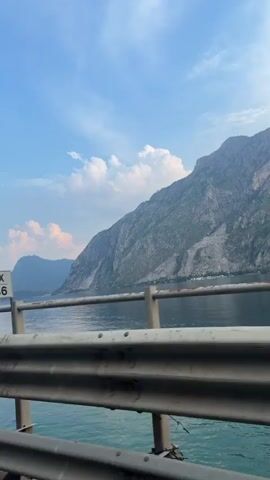 The video shows a view from a moving vehicle, likely a car, looking out over a body of water towards mountains. The water is a deep blue-green and appears calm. A guardrail, made of metal, is prominent in the foreground, obscuring part of the view. The mountains are rugged and covered in green vegetation. The sky is blue with scattered white clouds. The lighting suggests it is daytime, possibly late afternoon or early morning given the soft light. There are no people visible in the video. The camera is positioned low, close to the guardrail. The video is very short, approximately three seconds long, and the movement suggests the vehicle is traveling along a coastal road or a road adjacent to the body of water.