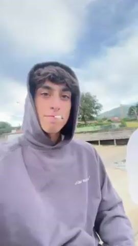The video shows two young men sitting on a concrete structure overlooking a body of water. The first man, closer to the camera, wears a gray hooded sweatshirt and has a cigarette in his mouth. The second man sits beside him in a white long-sleeved shirt and purple pants. The camera then pans to show a wide view of the lake, with a cloudy sky overhead and the opposite shoreline visible in the distance.