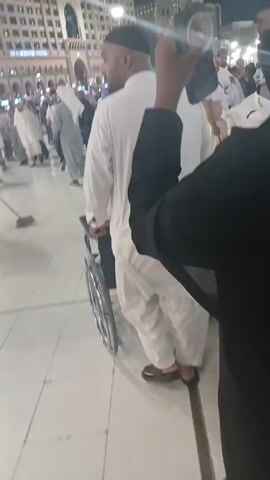 The video captures a street scene at night with a crowd of people, many of whom are wearing white robes, suggesting a religious gathering or pilgrimage. The crowd moves along a wide, paved street. A large, modern building with many windows looms in the background. The perspective is from someone moving with the crowd, occasionally showing the back of a man wearing a black cap. The video is somewhat shaky and the focus varies. The lighting is bright, likely from streetlights or building illumination. Some individuals are taking photos or videos with their phones. A person wearing a black garment is briefly seen holding up a phone.