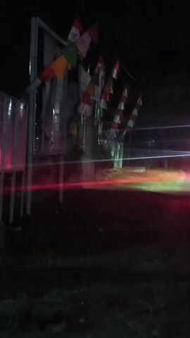 This is a very dark, low-light video. In the foreground, the ground appears to be covered in dry leaves and grass. A vertical white pole with some metallic structures is visible on the left, with what look like red lights reflecting off them. To the right of this pole, there is a darker vertical structure, possibly a wooden pole or a beam, from which a string of small triangular flags is hanging. The flags are in various colors, including red, white, green, and orange. Some of the flags appear to be in a triangular shape, while others are more like pennants.

Further back, there is a blurry background suggesting a road or open area. Streaks of red and green light are visible, indicating movement, likely from vehicles passing by. The overall impression is a nighttime scene, possibly in a rural or semi-urban setting, with some festive decorations. The video is shaky and the focus is not sharp due to the low light conditions.