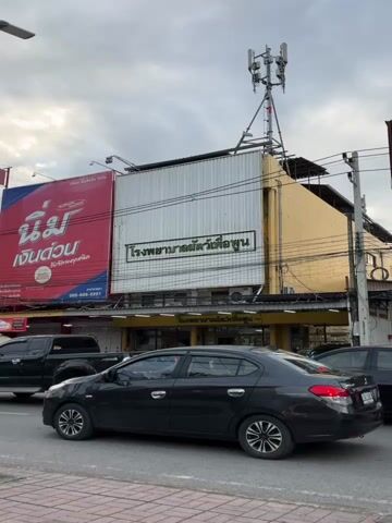 This video shows a street scene in what appears to be Thailand. A black sedan is driving in the foreground, and a white scooter passes it. In the background, there is a building with a large sign in Thai script that reads "โรงพยาบาลสัตว์ เล็ก ทวีโชค". Above the building, a cell phone tower is visible. There are other vehicles and buildings in the background, and the sky is overcast. The video appears to have been filmed from a moving vehicle.