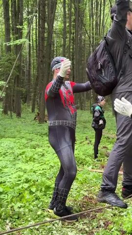 In a verdant forest setting, a group of individuals participates in an outdoor activity involving ropes. In the foreground, a male figure wearing a black and red Spider-Man full-body suit, a black and white bandana, and white gloves stands on the left, facing right, with his arms extended forward, palms open, appearing to support or guide another male figure to his right. This second male figure, wearing a dark jacket, dark pants, a black baseball cap, and white gloves, with a black backpack, is attempting to traverse a low-strung horizontal rope, using two vertical ropes hanging from above for balance and support. Only his left foot is clearly visible on the rope, while his right foot is slightly lifted. Behind him, another person, largely obscured, provides support. In the background, a female figure in dark clothing and a bandana stands observing, while a male figure in a dark shirt and blue cap sits on the ground further to the right, also observing. The ground is covered with green foliage, and tall trees form the backdrop.