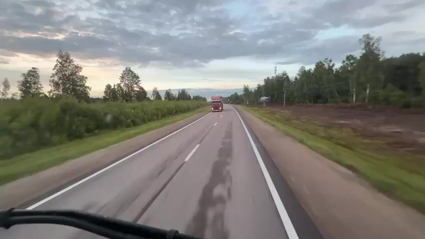 The video is a first-person view from a vehicle driving on a two-lane asphalt road. The road is wet, indicating recent rain. On the left side of the road, there are green bushes and trees, and on the right side, there are more trees and a cleared patch of land with some dirt. The sky is cloudy with hints of sunset colors. There is a red truck in the distance traveling in the opposite direction. Further down the road, a large blue semi-truck is passing by. The vehicle continues to drive forward, with the road stretching out ahead.