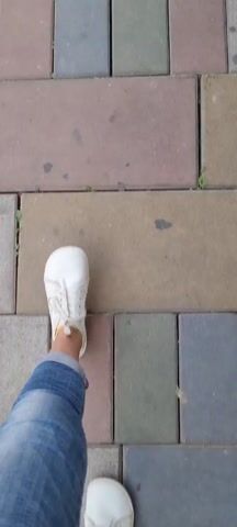 A person wearing white shoes and blue jeans walks on a paved surface made of rectangular bricks of varying colors including gray, brown, green, and blue. The camera focuses on the person's feet and the paved surface as they walk.