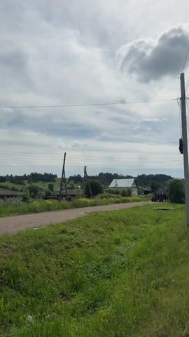 A medium shot captures a rural road in Lukintsy, Russia, on an overcast but bright day. The scene is dominated by lush green grass in the foreground and along the roadside. A paved road stretches from the left of the frame towards the right, curving slightly.

A tractor, possibly a MTZ (Belarus) model, with a trailer attached, drives from left to right along the road. In the background, scattered houses with light-colored roofs are visible amidst rolling hills covered with trees and greenery. Power lines span across the upper part of the frame, connecting several utility poles, one prominently on the right. Another wooden utility pole stands further back on the left side of the road. The sky is partially cloudy with a mix of white and grey clouds. No people or visible text are present in the video.