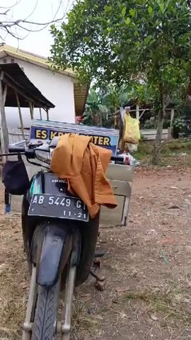 The video opens with a close-up of a motorcycle with a sidecar containing a cooler and advertising for "ES KRIM ROTI". A brown jacket is draped over the cooler. The motorcycle is parked on a dirt path in front of a small building with a covered porch. The camera pans to the left, revealing a covered structure with benches and several people standing or sitting. The environment appears to be outdoors, with dry grass and some trees. In the background, there are more buildings and some signs of human activity, including what looks like a schoolyard with children in uniform. The video then shows a person on a motorcycle with a similar cooler, interacting with others under the covered structure. The overall scene suggests a street vendor selling ice cream in a village or school setting.