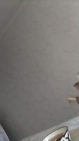 A handheld video begins with a close-up of a bowl of liquid, possibly tea, on a patterned surface. The camera then pans to the left, revealing a white mug with a black handle, also on the patterned surface. In the background, a light-colored wall with a subtle textured pattern is visible. A large object draped with a light-colored cloth, featuring a pattern of small, circular shapes and stripes, occupies the middle ground. To the left of this draped object, a doorway is partially visible, with a door ajar, revealing a dark interior. The floor is covered with a patterned carpet. The lighting suggests an indoor setting, possibly during the daytime. The overall mood is domestic and unremarkable. There is no visible text in the video. The camera movement is shaky, indicating it is being held by hand. The video's timestamp begins at 00:00 and ends at 00:05.