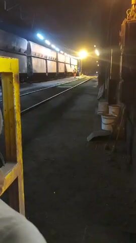 The video shows a nighttime scene at what appears to be a train yard or industrial facility. A long train of grey railcars is parked on tracks. Bright artificial lights illuminate the scene from overhead, casting long shadows. A person wearing an orange safety vest is visible near the train. The ground is covered in dark material, possibly coal dust or dirt. To the right of the tracks, there are several white buckets and some tools leaning against a dark structure. A yellow metal barrier is in the foreground on the left. The camera angle suggests the viewer is positioned at a slightly elevated vantage point, looking down the length of the train.