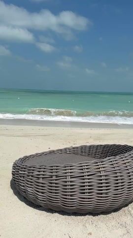 The scene is a sun-drenched beach featuring a large, round, gray woven rattan lounger in the foreground. Behind the lounger, the frame shows a calm, turquoise sea meeting the white sand beach. Gentle waves are visible along the shoreline. The sky is a clear blue, dotted with a few fluffy white clouds. The scene appears peaceful and serene. There are no people visible in the shot.