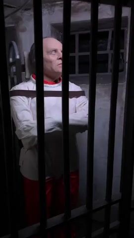 A person dressed as Hannibal Lecter stands behind bars in a dark room. The person wears a white straitjacket and red pants. The background is a dark, possibly padded cell. The image fades to black, and then a loud crashing sound is heard.