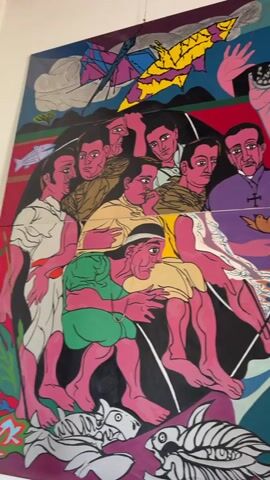 The video shows a wall mural comprised of several individual panels. The style is colorful and folk art-like, with simplified depictions of people and elements. In one panel, a group of men are gathered, with a priest figure wearing a purple robe with a cross and a dove on it. Below, there's a figure in a green garment and a school of white fish. Another panel features a figure with pink skin in brown clothing, holding a dark object with a white symbol. A third panel depicts a pair of figures, one with their hand raised. The artwork features bright colors, including blues, greens, purples, yellows, and reds. The backdrop includes abstract shapes, boats, and birds.